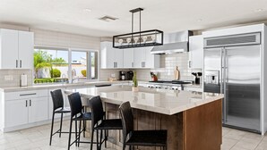 A modern kitchen with white cabinetry, a large island with bar stools, a stainless steel refrigerator, and a view of the outdoors.