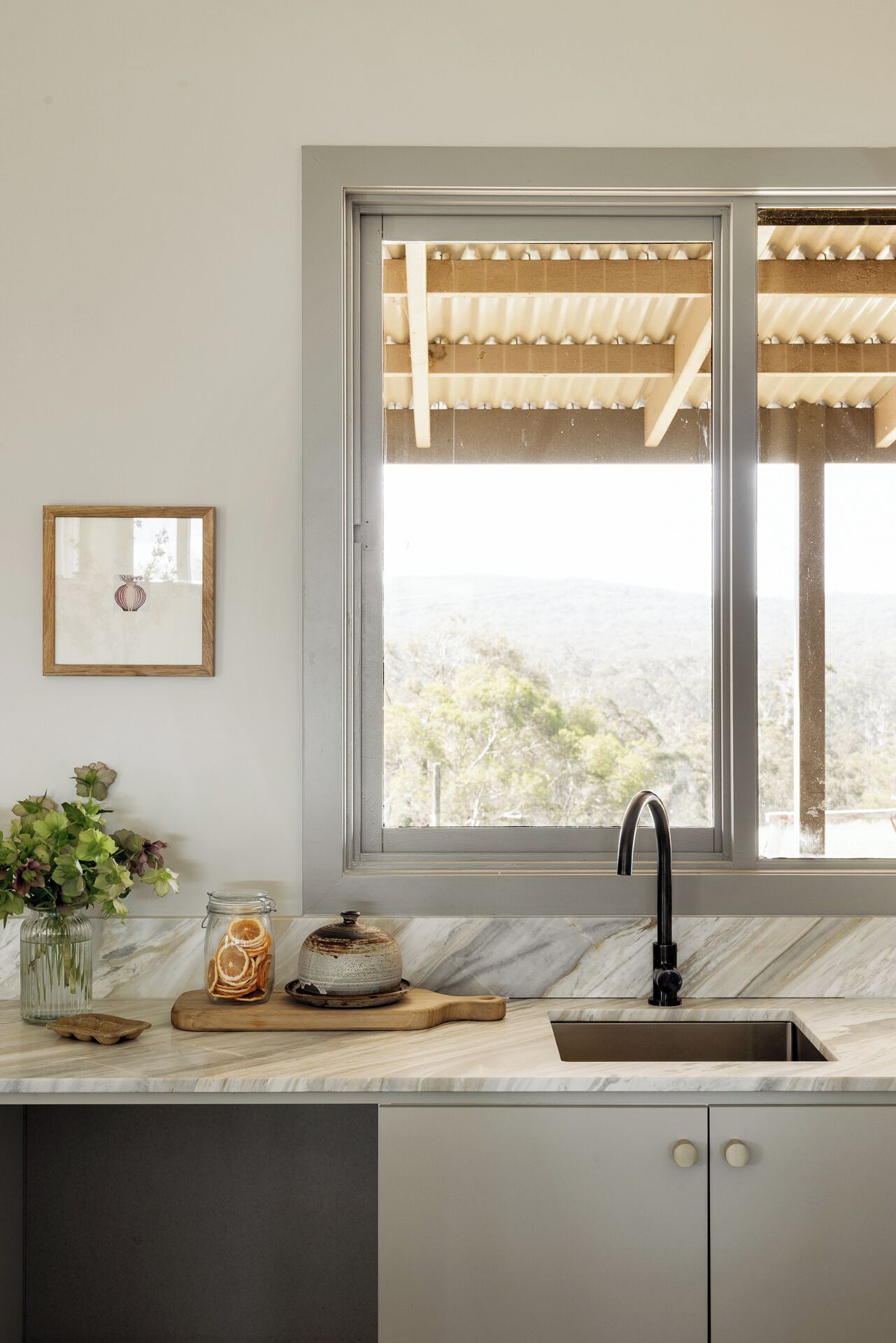 A kitchen with a large window, a sink, a vase with green leaves, and a jar of dried orange slices.