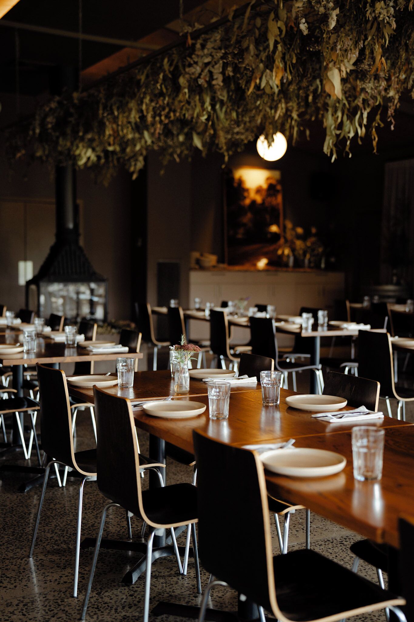A restaurant with long wooden tables, black chairs, and a hanging plant decoration.