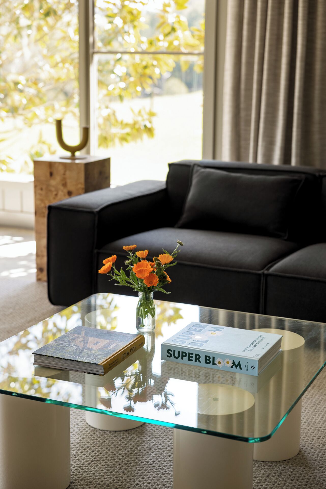 A modern living room with a glass coffee table, a black sofa, and a vase with orange flowers.