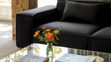 A modern living room with a glass coffee table, a black sofa, and a vase with orange flowers.
