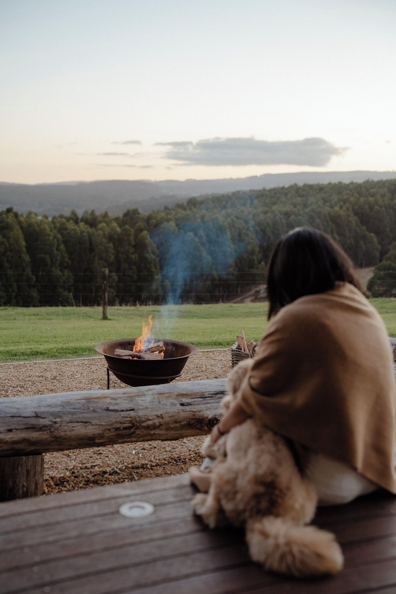 Une personne assise près d’un feu de camp, emmitouflée dans une couverture, et qui contemple un paysage boisé.