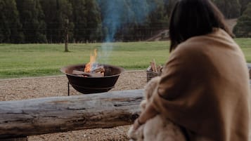 Une personne assise près d’un feu de camp, emmitouflée dans une couverture, et qui contemple un paysage boisé.