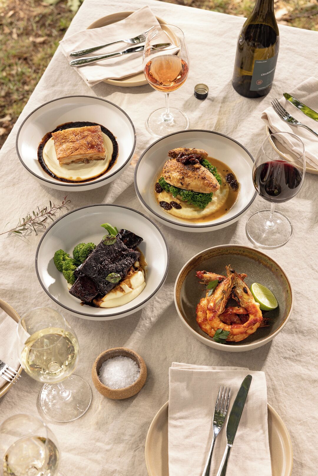 A table set with various dishes, including a piece of meat, seafood, and a dessert, accompanied by wine glasses and cutlery.