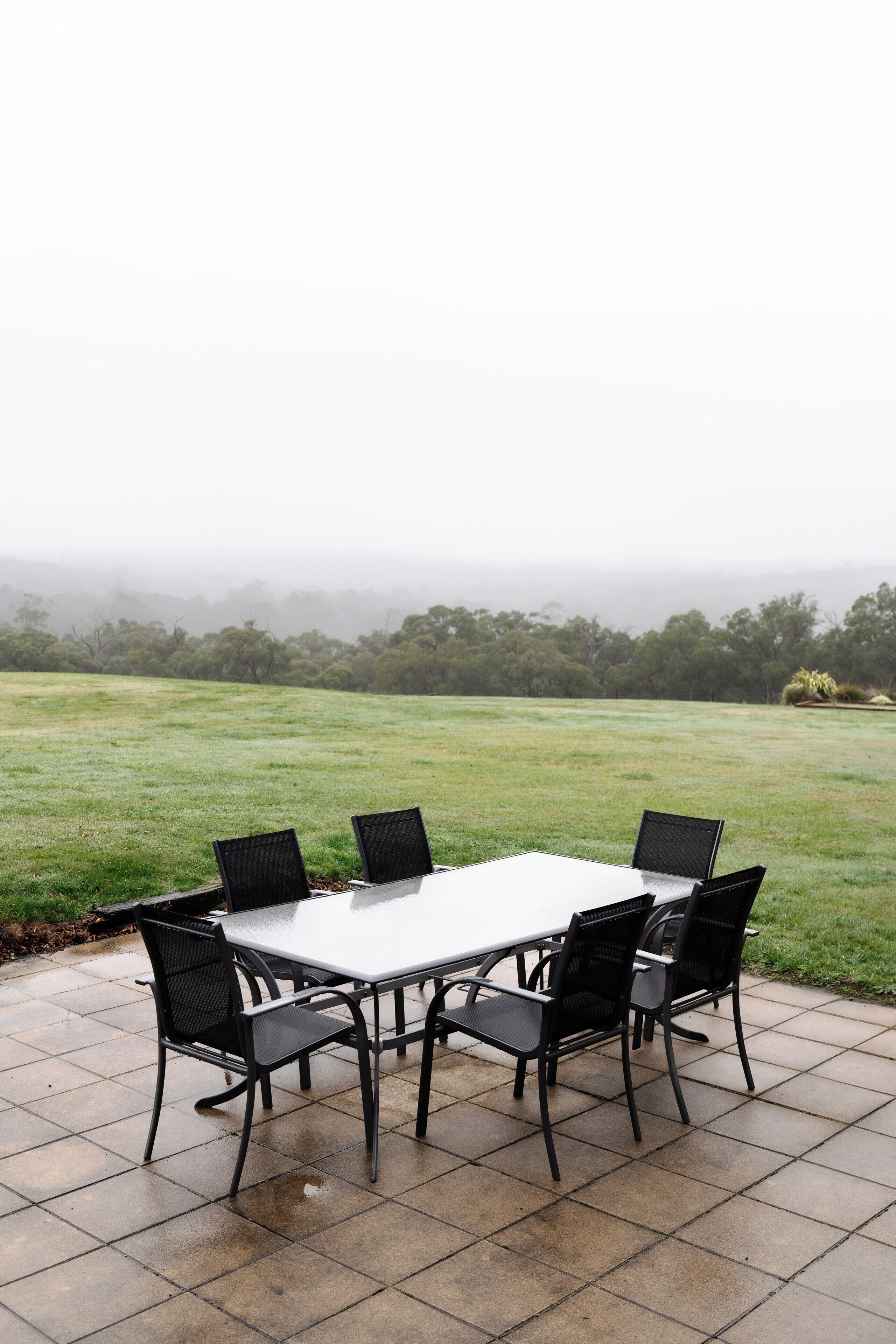 A patio table with four chairs, a white table, and a view of a grassy field and trees in the distance.