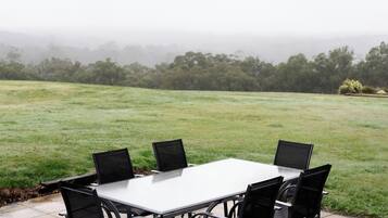 A patio table with four chairs, a white table, and a view of a grassy field and trees in the distance.