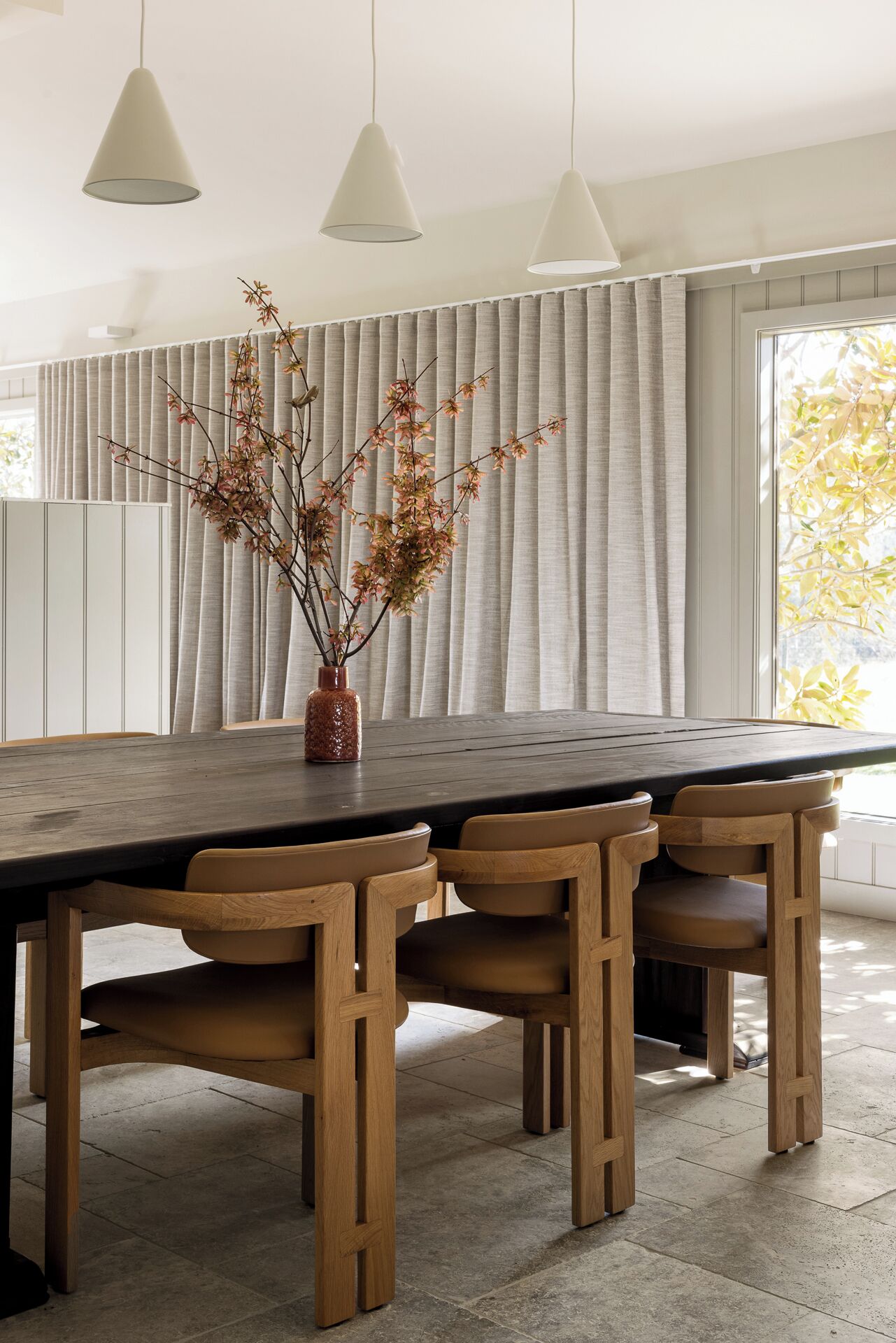 Een eetgedeelte met een lange houten tafel, zes stoelen en een vaas met takken.