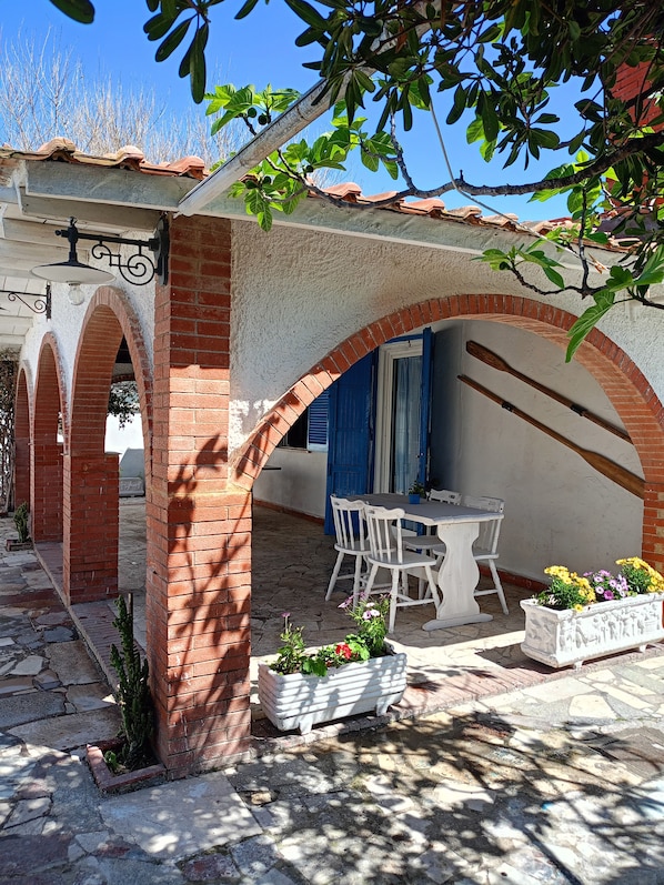 A covered outdoor seating area with a table and chairs, surrounded by brick arches and potted plants.