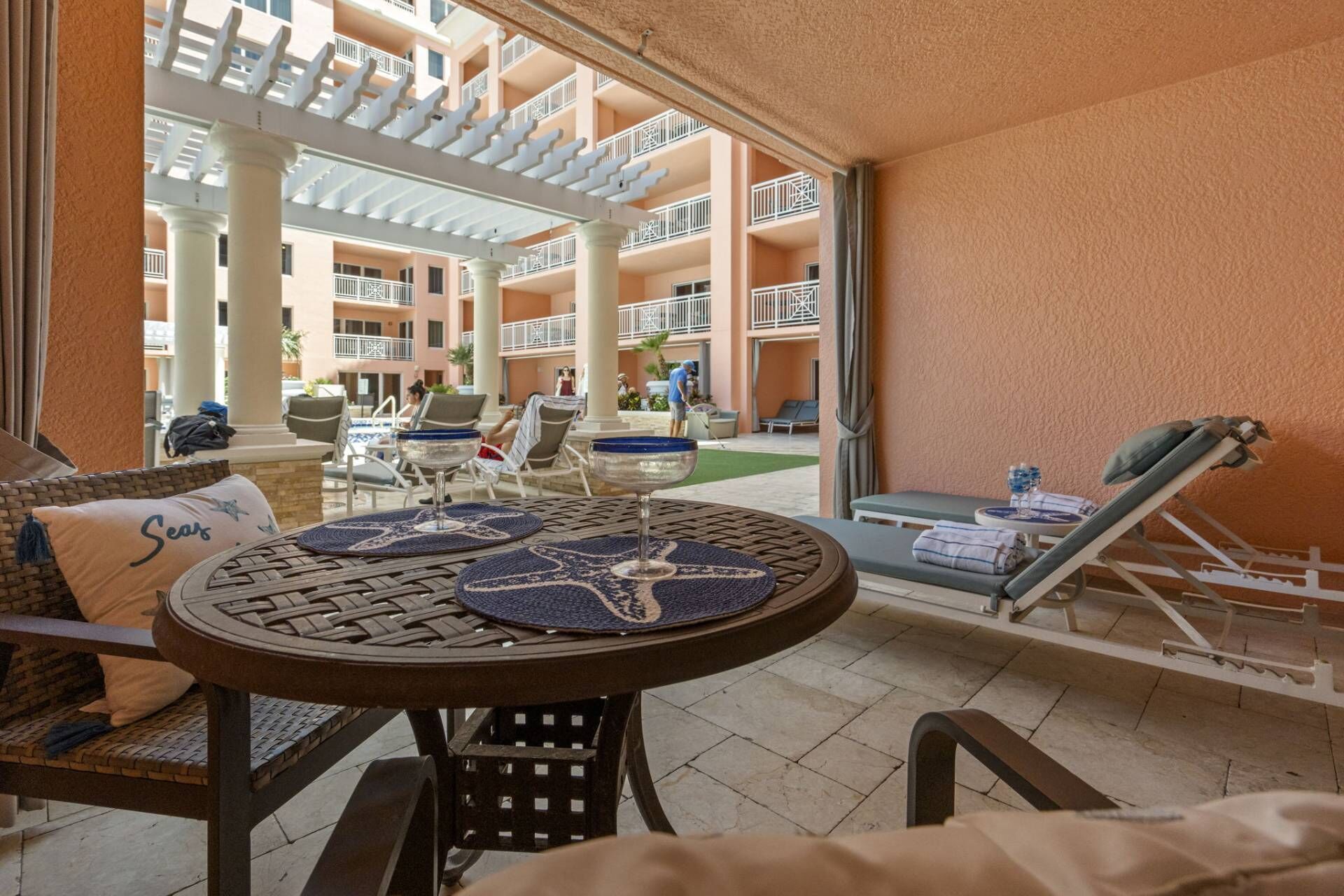 A sunlit outdoor seating area with a round table, lounge chairs, and a view of a multi-story building with balconies.