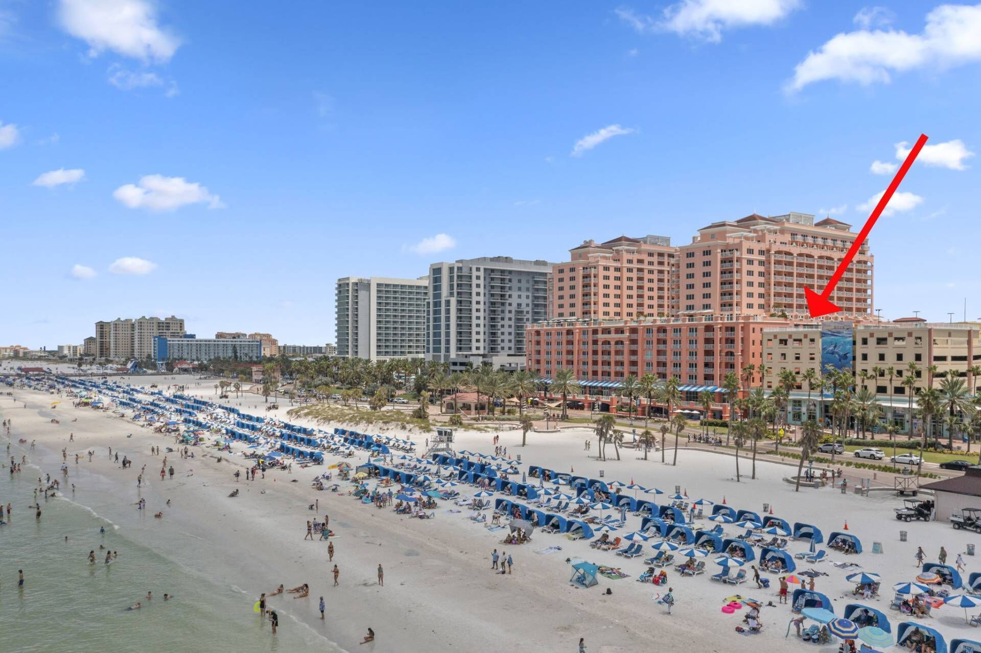 Un resort en la playa con varios edificios altos, una playa de arena con numerosos sombrillas y sillas de descanso, y un cielo azul despejado.