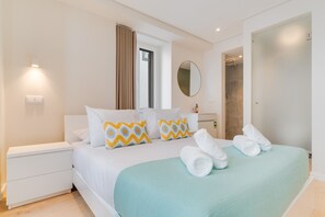 Quarto de hotel moderno com cama, mesas de cabeceira brancas, banheiro e um espelho grande.