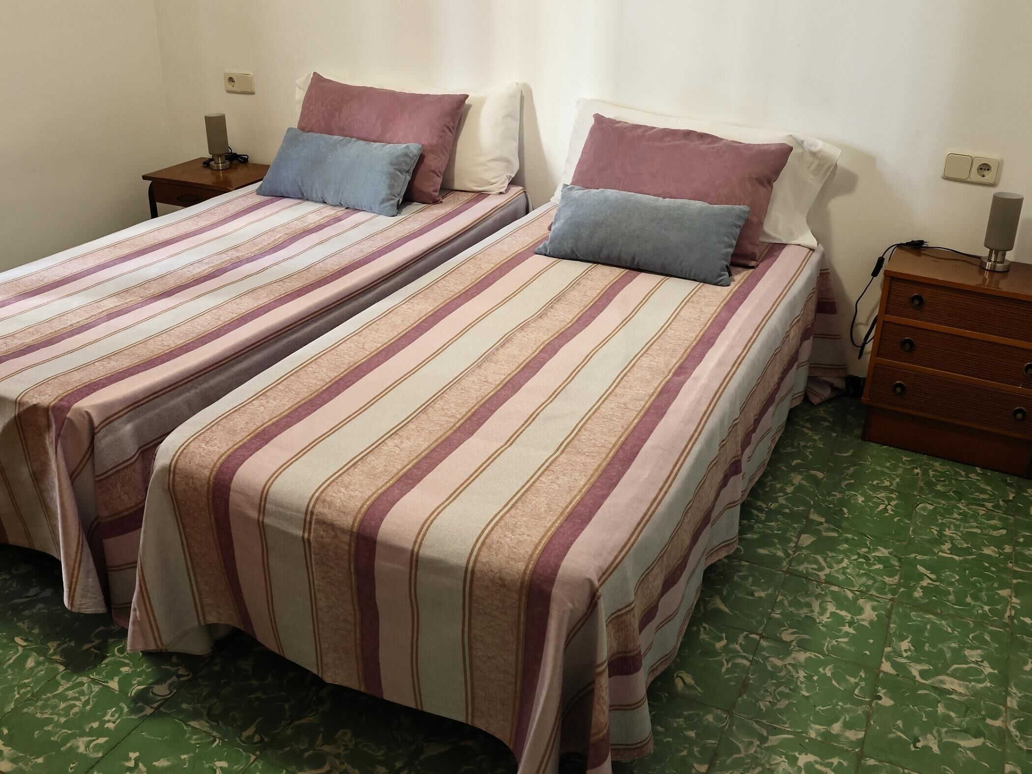 Two single beds with striped bedding, a wooden nightstand with a lamp, and a green patterned floor.