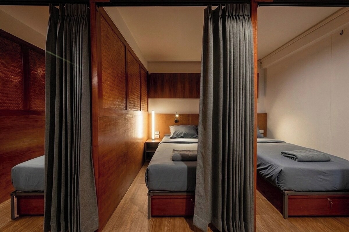 A hotel room with a wooden floor, two beds with grey bedding, a nightstand with a lamp, and a window with curtains.