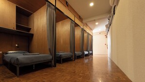 Egy, a szobához vezető, fa padlóval ellátott folyosó, ahol négy ágy található, mindegyik szürke matraccal és fafejtámlával van felszerelve.