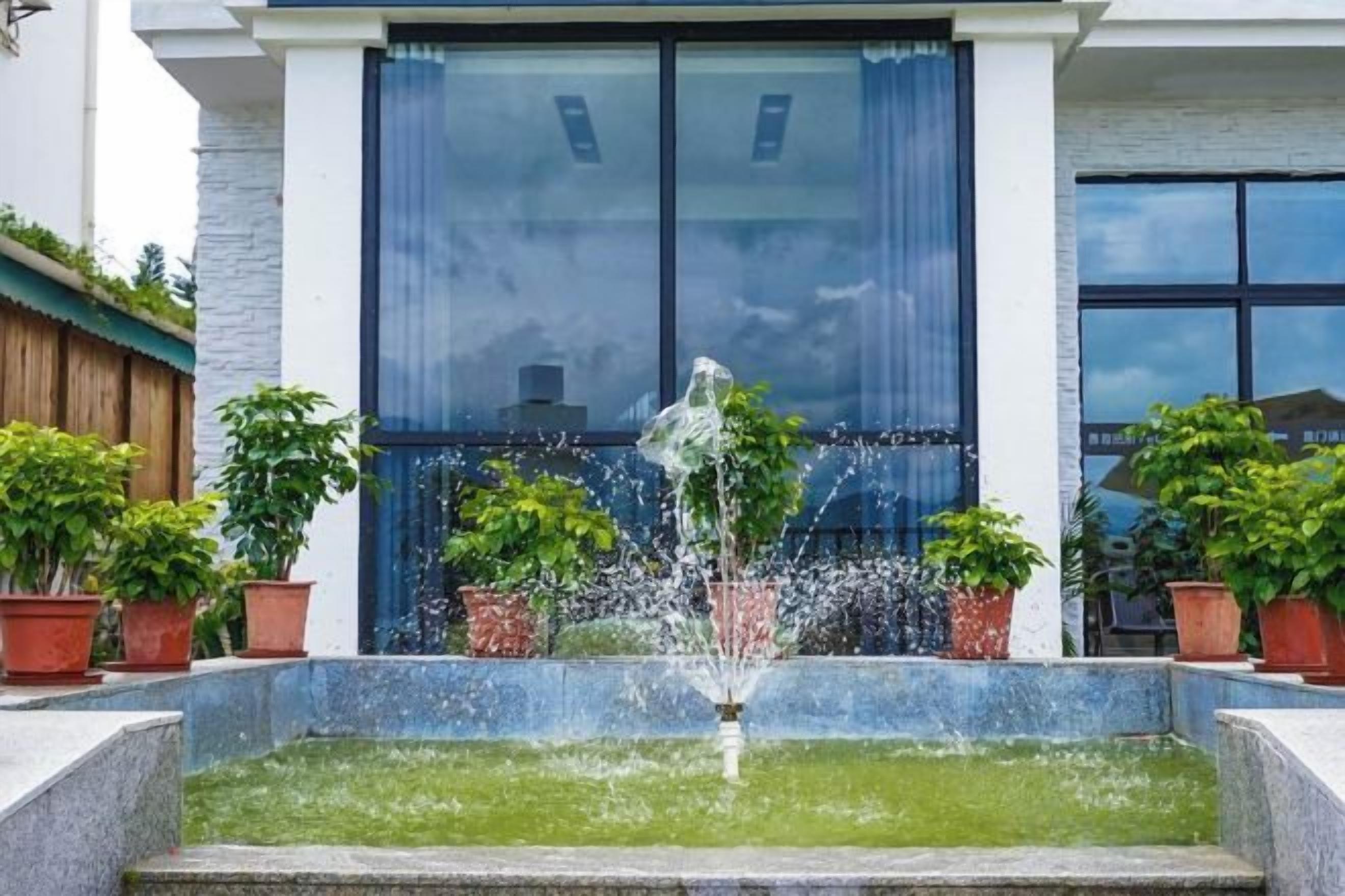 Una fuente con chorros de agua en una pileta rectangular, rodeada de plantas en macetas, frente a un edificio con grandes ventanales y fachada de piedra.
