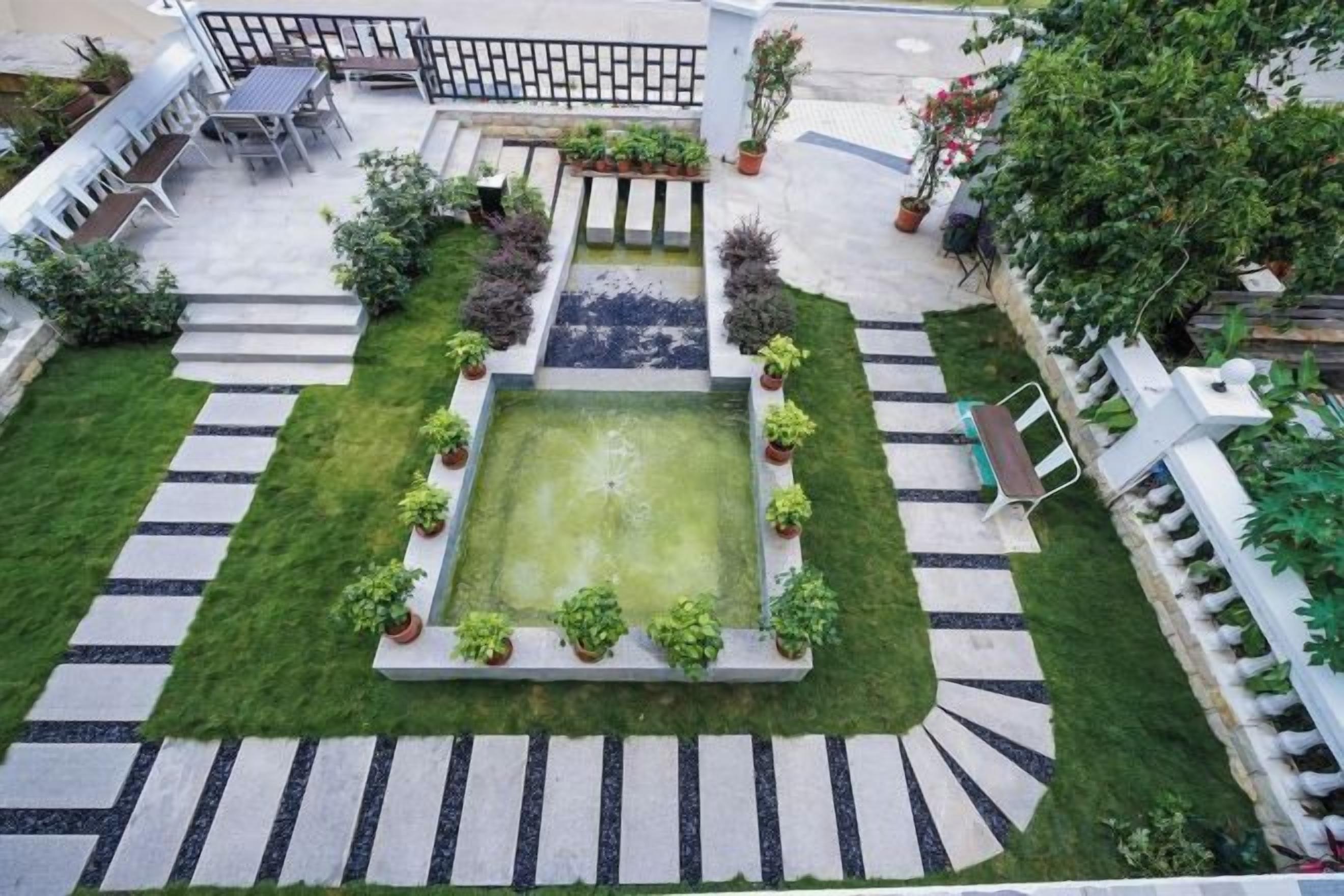 A courtyard with a central square pool, surrounded by a garden with various plants and a seating area with tables and chairs.