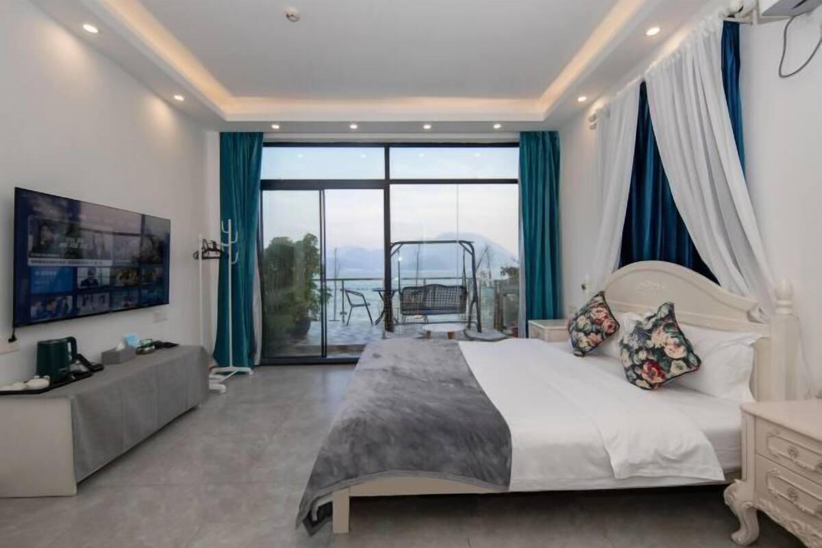 Un dormitorio con una cama grande, televisión, balcón con una silla y cortinas.