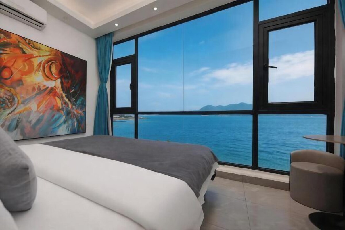 A bedroom with a large bed, a painting, a view of the sea, and a chair.