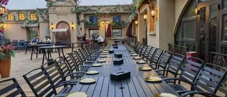 Un área de comedor al aire libre con mesas y sillas largas, rodeada por un edificio con ventanas de arco y asientos exteriores.