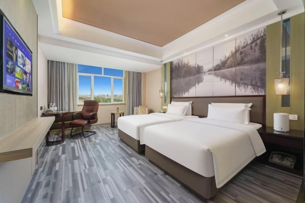 Une chambre d’hôtel avec un lit, un bureau, une chaise, une fenêtre donnant sur un paysage agréable et un tableau au mur.