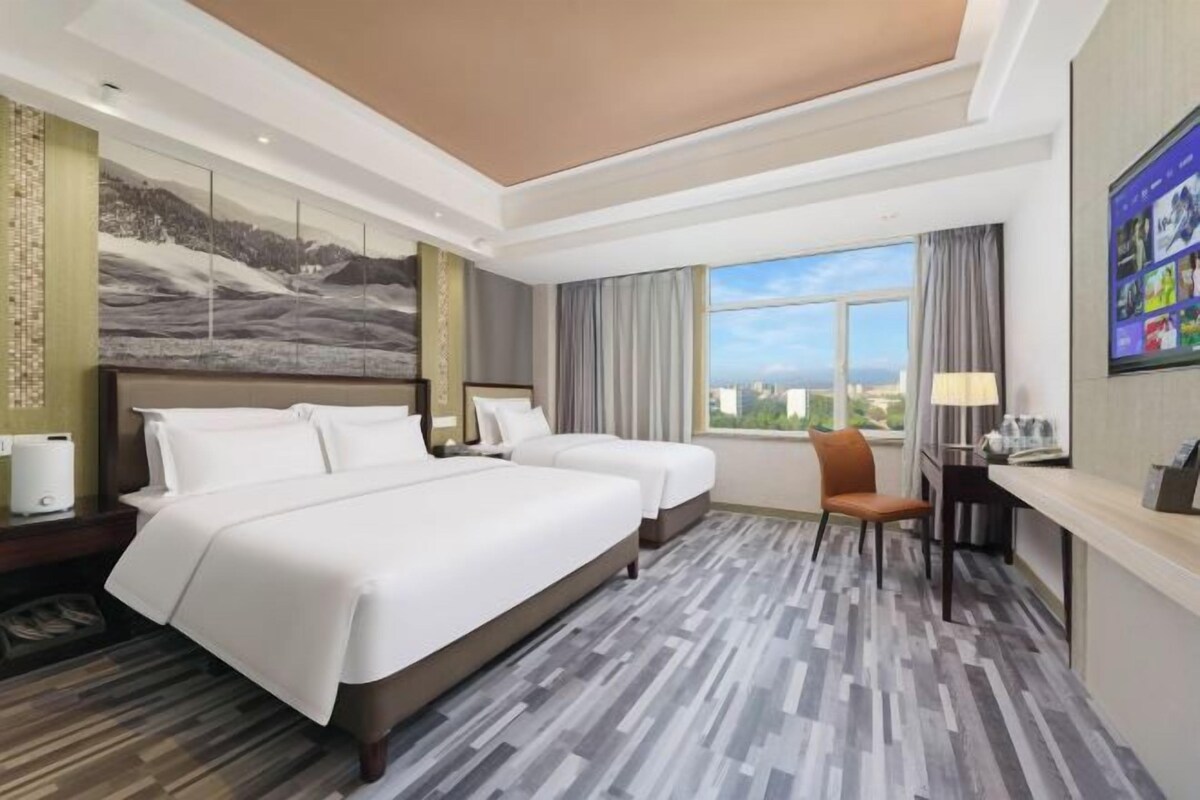Une chambre d’hôtel avec deux lits, un bureau, une chaise et une grande fenêtre donnant sur la ville.