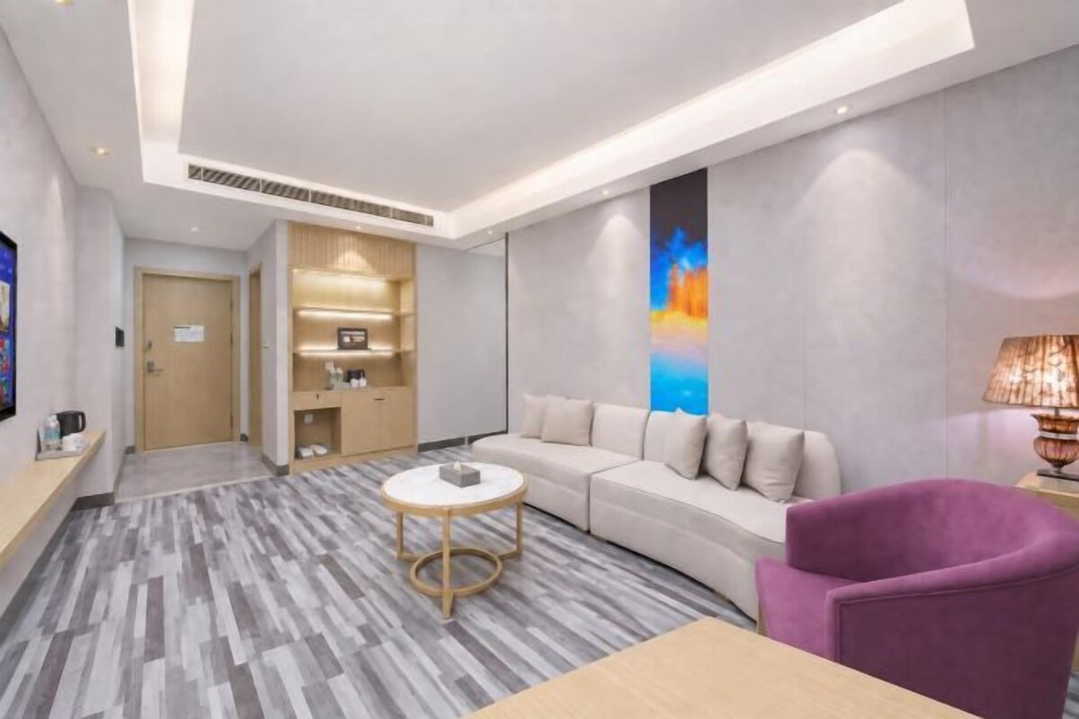 A modern hotel room with a sofa, a purple armchair, a small round table, and a bookshelf.