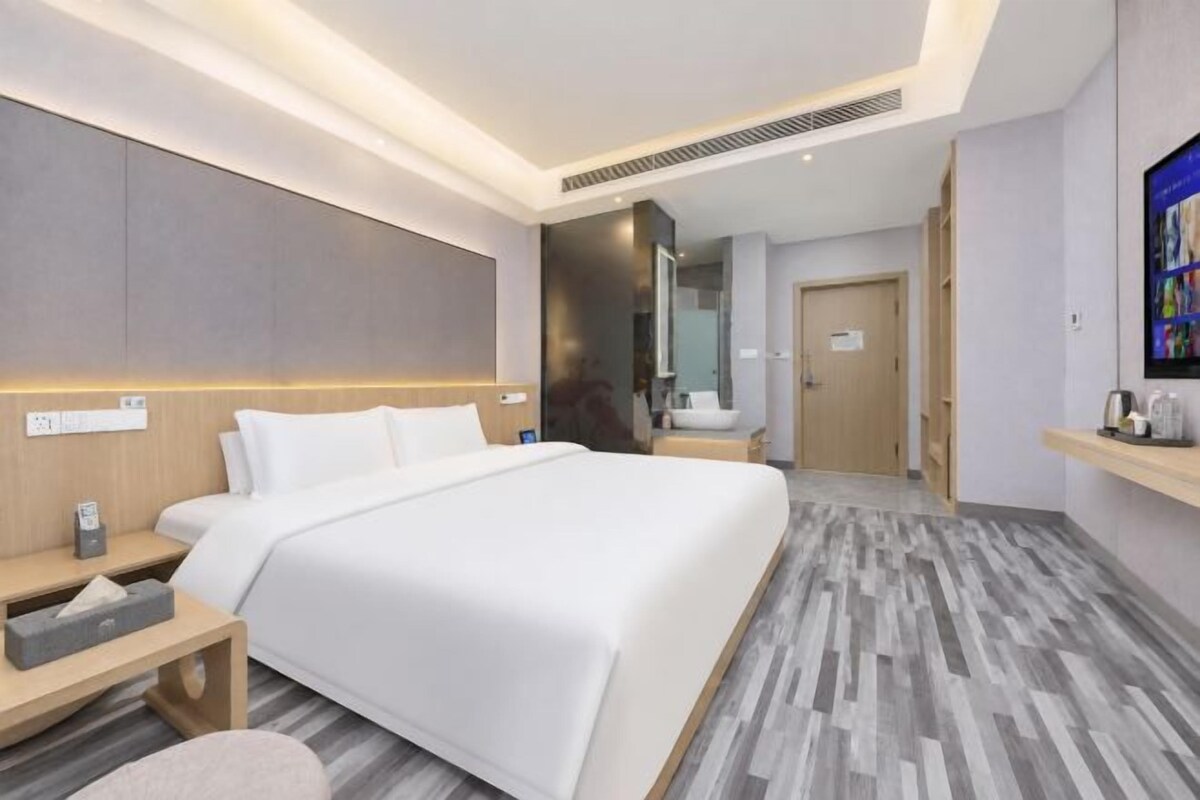 A modern hotel room with a large bed, bedside tables, a nightstand, a mirror, and a small seating area.