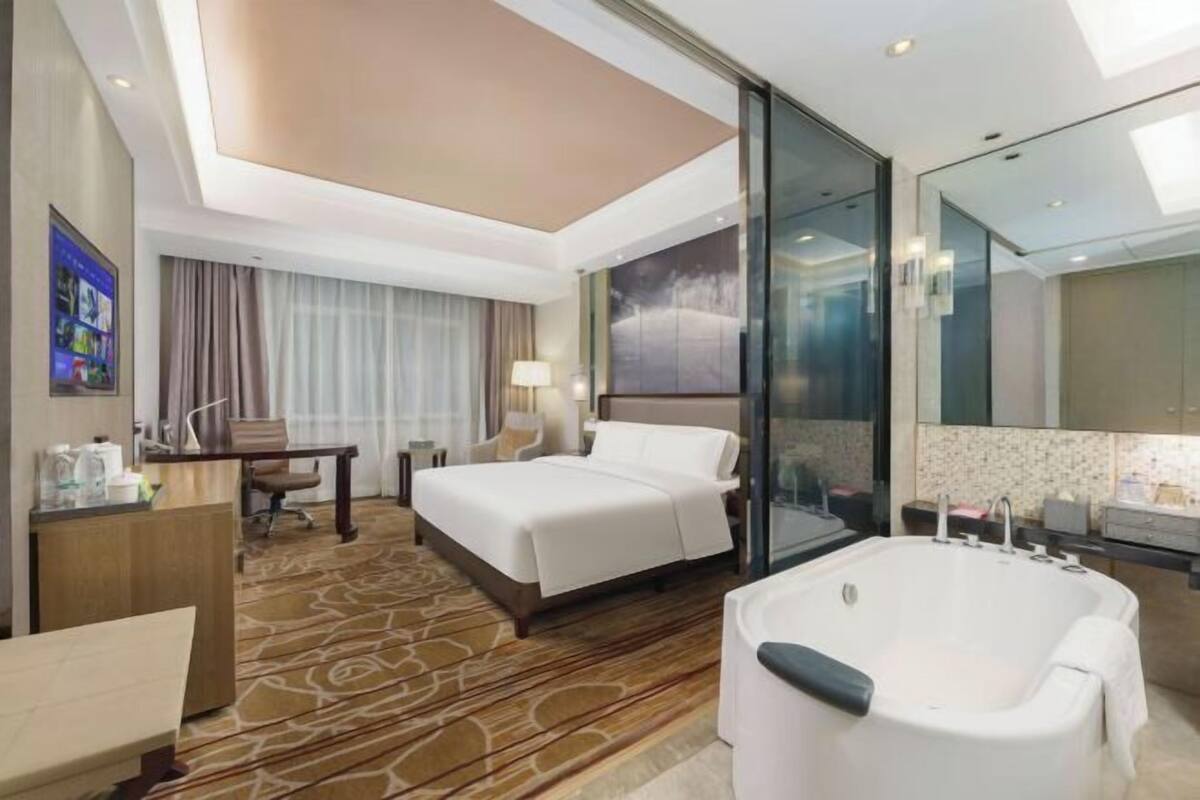 Une chambre d’hôtel avec un grand lit, un coin salon avec un fauteuil, un bureau avec une lampe, une grande fenêtre avec des rideaux, une salle de bain avec une baignoire et un grand miroir.