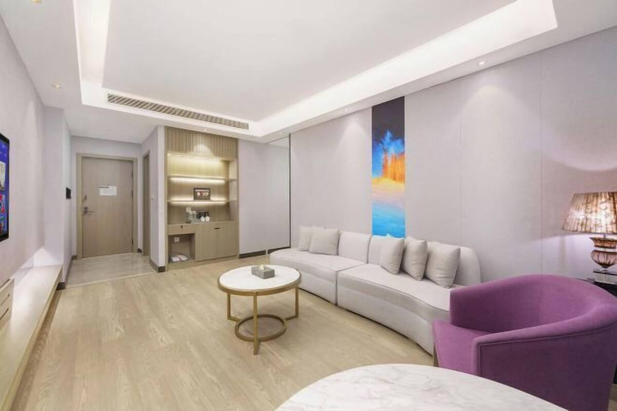 Une chambre d’hôtel moderne avec un canapé incurvé, un fauteuil violet, une petite table ronde et un grand tableau abstrait accroché au mur.