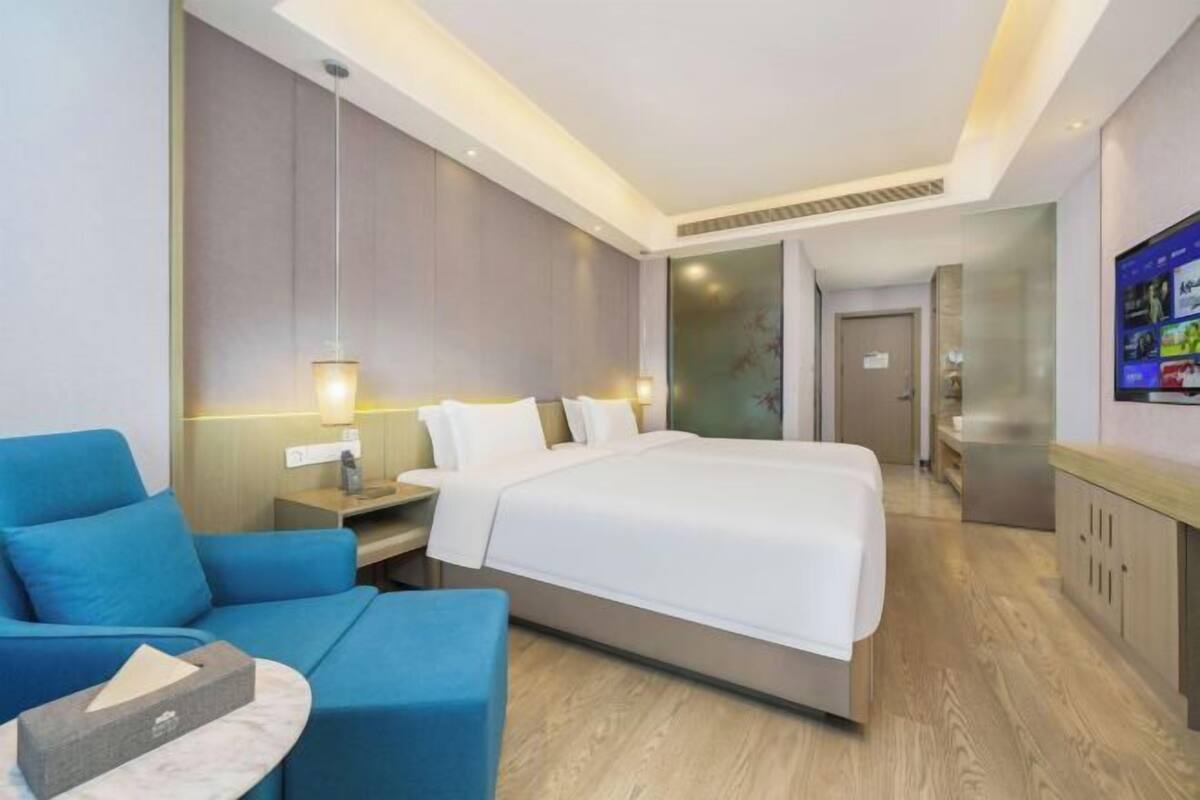 Une chambre d’hôtel moderne avec un grand lit, un fauteuil bleu, un bureau avec une lampe et un téléviseur fixé au mur.