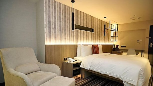 Quarto de hotel com uma cama grande, área de estar com uma poltrona, mesa de cabeceira com telefone e luminária fixada na parede.