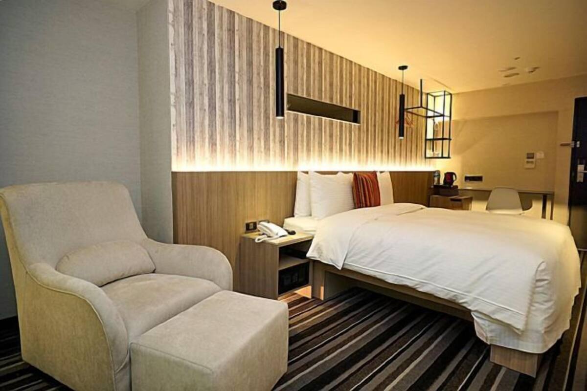 A hotel room with a large bed, a sitting area with a chair, a nightstand with a phone, and a wall-mounted light fixture.