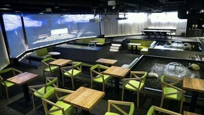 Restaurante com várias mesas e cadeiras, um pequeno elemento aquático e grandes telas exibindo conteúdo.