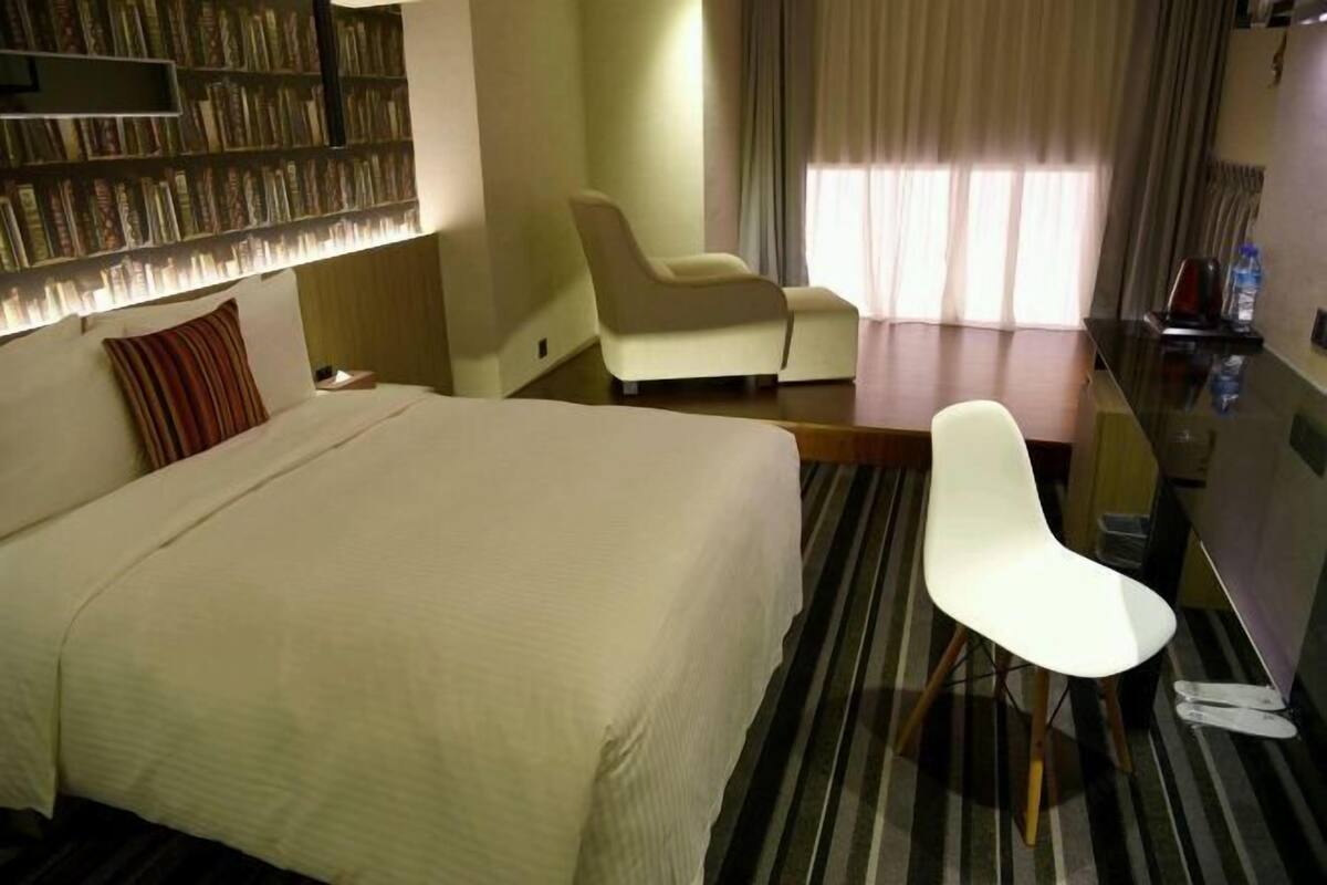 A hotel room with a bed, a desk, a chair, a bookshelf, and a window with curtains.