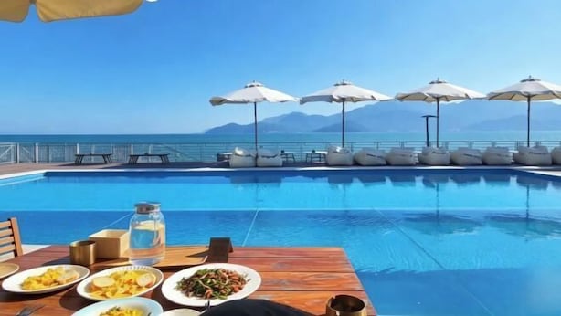 Área de refeições à beira da piscina com uma mesa posta para dois, vista desobstruída do oceano e vários guarda-sóis.