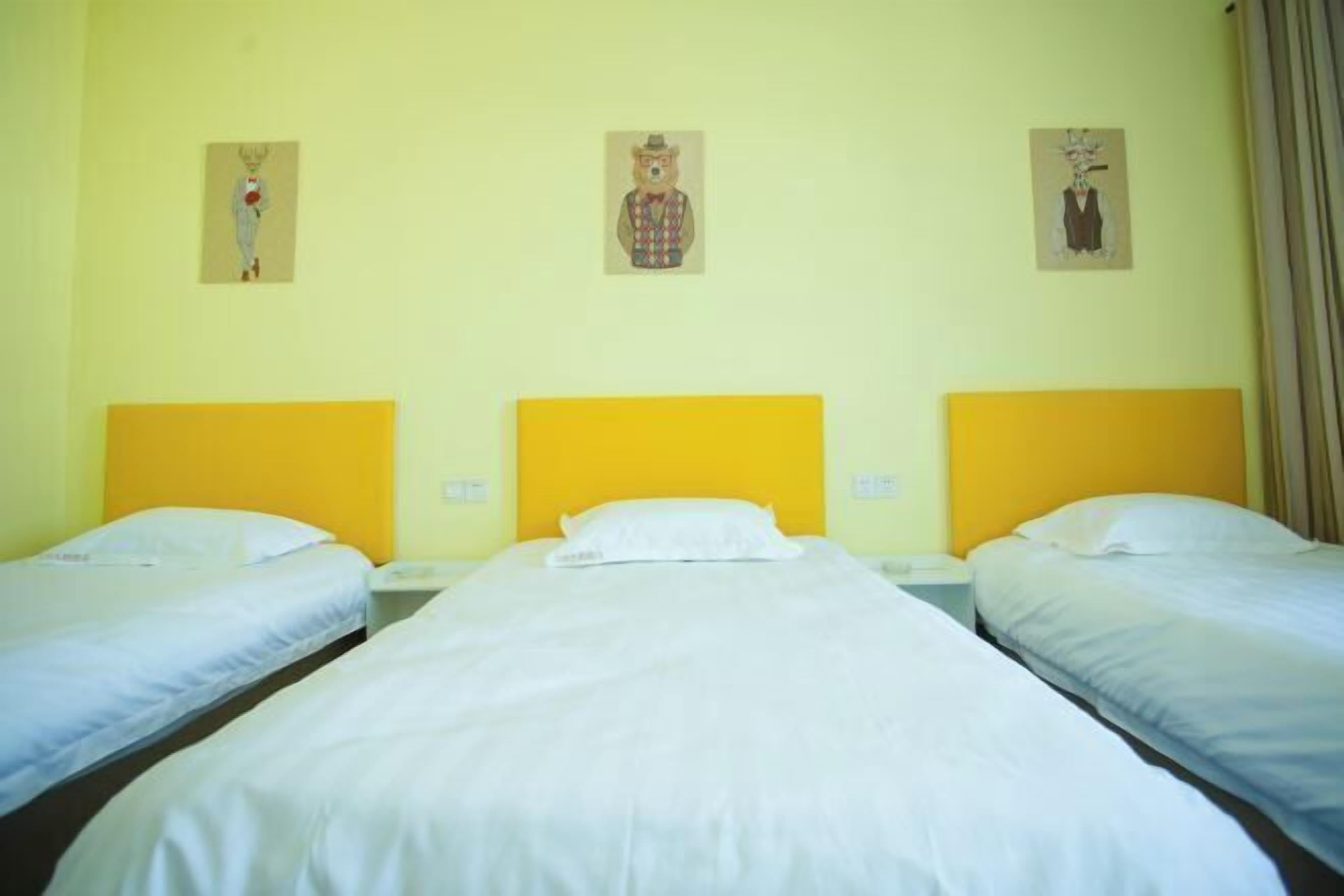 A hotel room with three beds, each with a yellow headboard, white bedding, and a small painting above each bed.