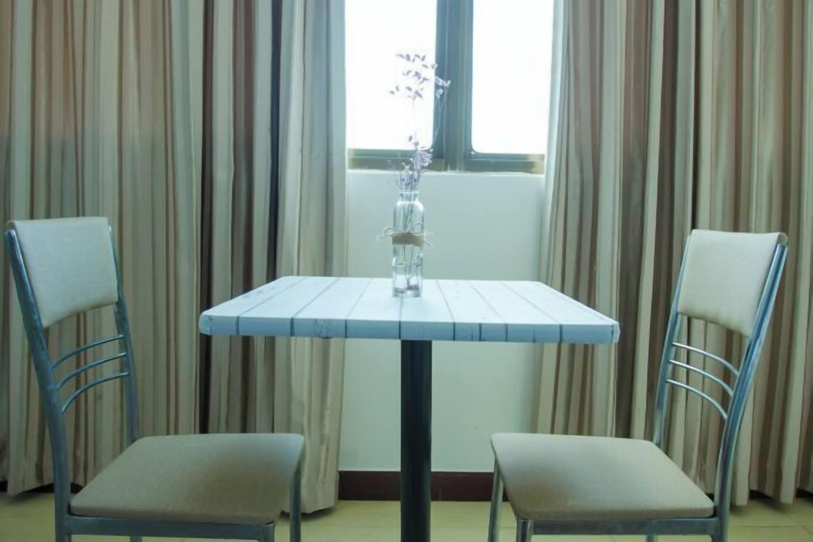 Una habitación con una pequeña mesa y dos sillas, una ventana con cortinas y un jarrón con flores en el alféizar.