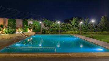 A nighttime swimming pool with a metal ladder, surrounded by lush greenery and illuminated by street lamps.