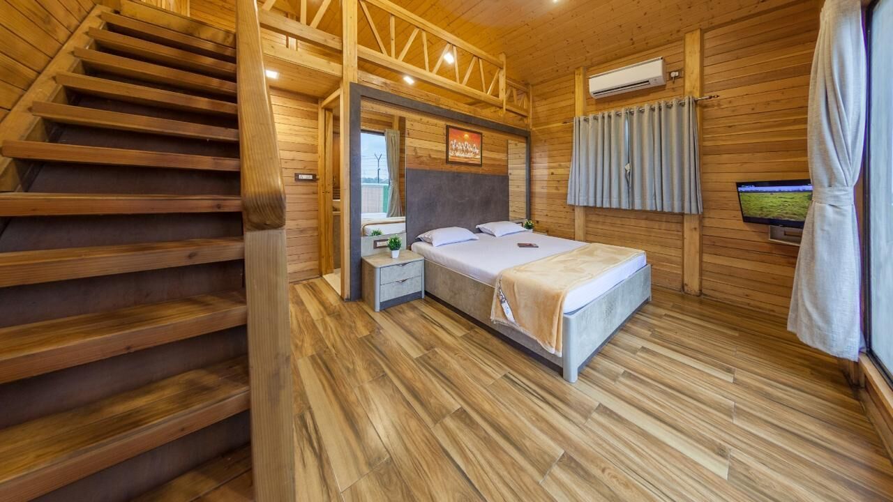 A wooden cabin bedroom with a bed, nightstands, a window with curtains, a TV, and a staircase.
