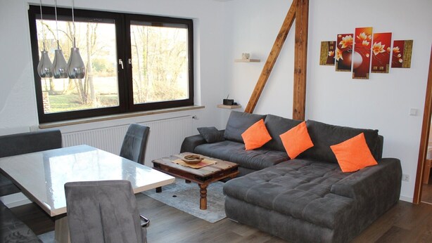 A modern living room with a grey sofa, orange cushions, a wooden coffee table, and a dining table.