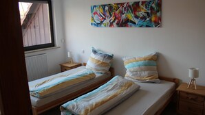 A room with two beds, a window, a radiator, a wooden nightstand, a lamp, and a colorful abstract painting on the wall.