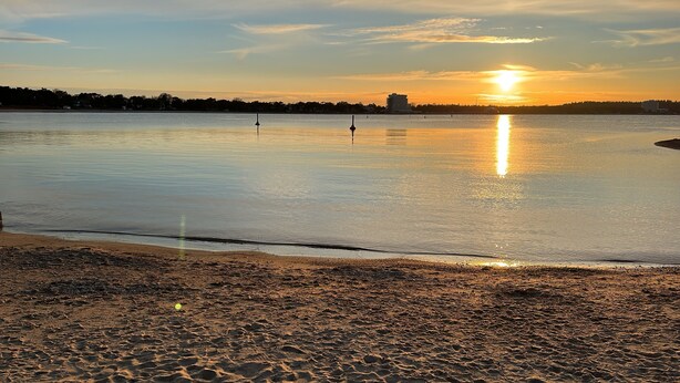A beach with a calm sea, a sunset, and a distant shoreline with buildings.
