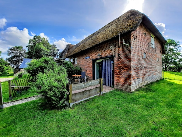 Un bâtiment en briques surmonté d’un toit de chaume, avec un petit espace extérieur aménagé pour s’asseoir et un chemin pavé menant à l’entrée.