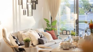 Ein modernes Wohnzimmer mit einer Couch, einem Esstisch mit Obstschale und einem großen Fenster mit Vorhängen.