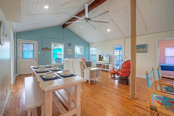 A spacious living area with a wooden floor, a dining table set with plates and cups, a ceiling fan, and a large window with a view of the outdoors.