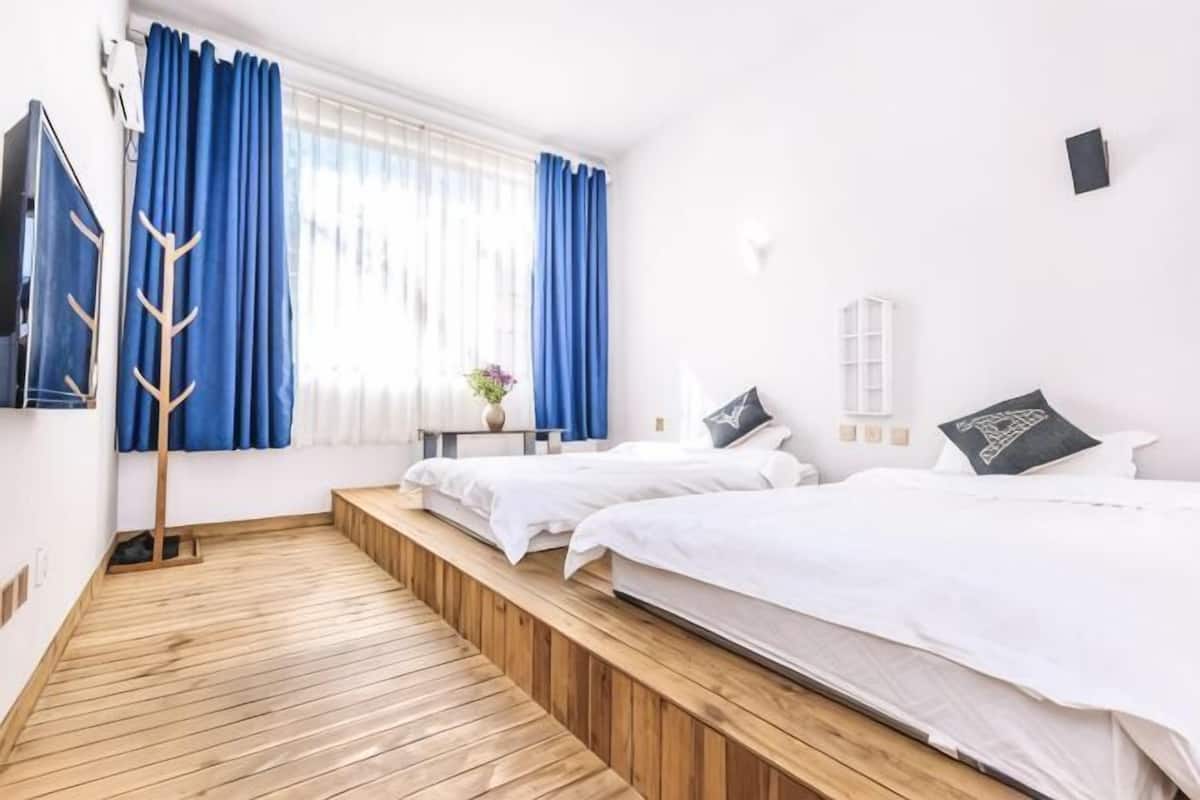 Une chambre d’hôtel avec deux lits, un plancher en bois, une fenêtre avec des rideaux bleus, un téléviseur fixé au mur et une petite table sur laquelle se trouve un vase.