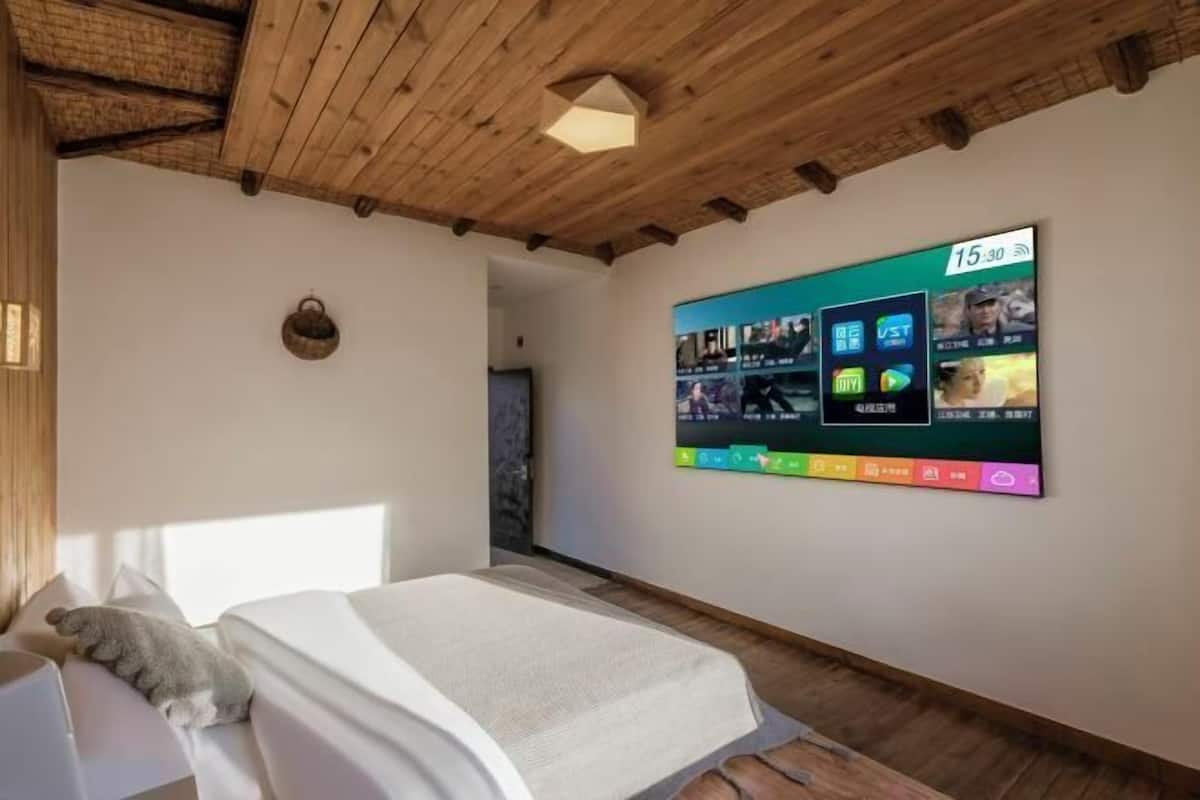 Une chambre d’hôtel moderne avec un plafond en bois, un grand écran numérique affichant diverses applications, un lit avec du linge de maison blanc et un petit panier fixé au mur.