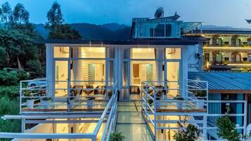 A modern two-story house with a glass facade, a covered outdoor area, and a mountain backdrop.