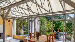 Un invernadero con una mesa y sillas de madera, una lámpara colgante y vistas de un jardín.