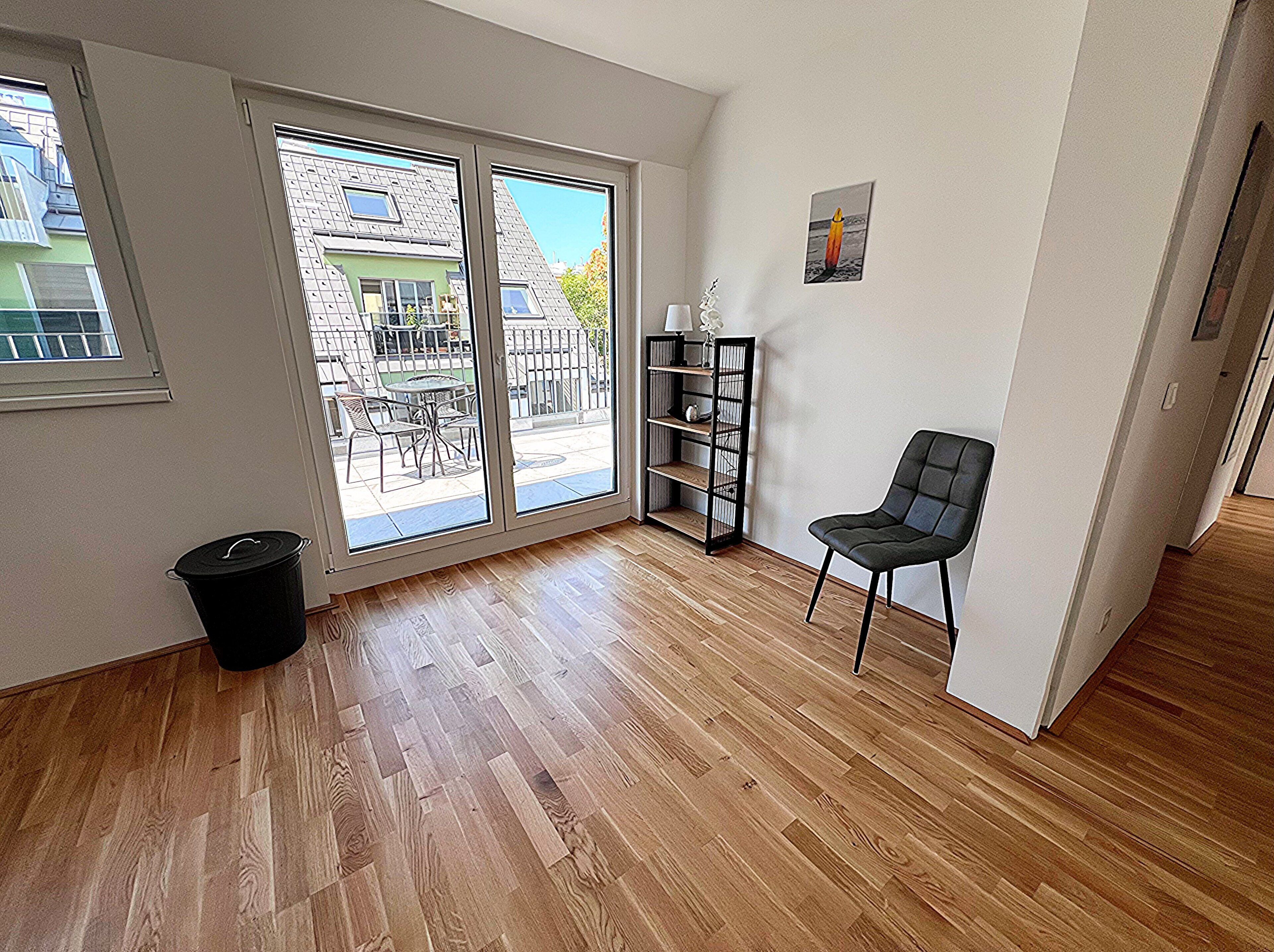 Une pièce dotée d’une porte-fenêtre coulissante donnant sur un balcon, d’une chaise noire, d’une bibliothèque et d’une poubelle.
